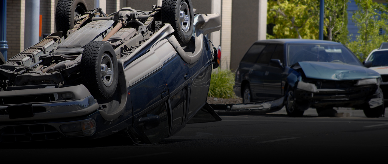 Photograph of a collision, a car with damage to the front hood and another overturned.